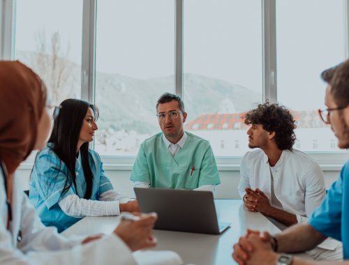 A group of multiethnic medical professionals including doctors, surgeons, and nurses are gathered in a hospital setting discussing patient care and using modern technology to address challenges in the medical field